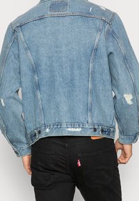 Person som bär en ljusblå sliten jeansjacka och svarta jeans med synliga bakfickor, stående mot en neutral bakgrund.