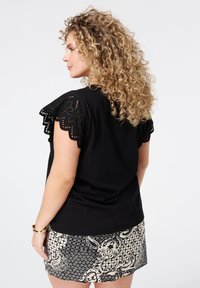 Haut noir avec des manches en dentelle à bords festonnés, associé à une jupe à motifs noir et beige, mettant en avant des textures et des motifs variés.