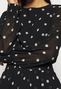 Svart blus med vita prickar, med genomskinliga ärmar, rund halsringning och rynkade manschetter. Tyget verkar lätt och strukturerat.