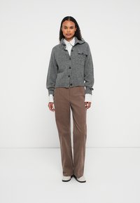 Kobieta w szarym swetrze zapinanym na guziki, w pasiastą koszulę, brązowe spodnie i białe buty, stojąca na tle gładkiej białej ściany.