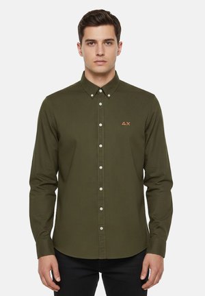 Uomo con capelli corti e scuri che indossa una camicia verde oliva a maniche lunghe abbottonata, con un piccolo logo arancione sul petto.