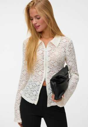 Femme aux longs cheveux blonds portant un haut blanc en dentelle zippé et un pantalon noir, tenant une pochette en cuir noire pliée.