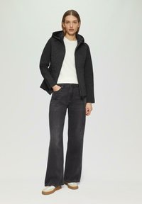 Zwarte gewatteerde hoodie met een gestructureerde voorkant, gecombineerd met donkere flare-jeans en lichte sneakers. De stof lijkt zacht en rekbaar.