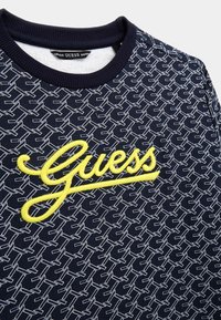Granatowy sweatshirt z białym geometrycznym wzorem, z haftowanym żółtym logo "Guess". Żebrowany kołnierz i miękka tekstura.