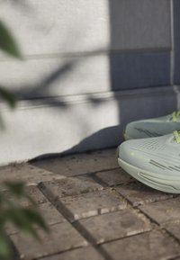 Light green running shoes feature a textured upper, reflective accents, and a cushioned sole, placed on a stone pavement with shadows.