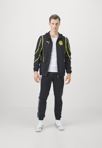Chaqueta deportiva negra con detalles amarillos y logo, combinada con pantalones de chándal negros. El modelo lleva una camiseta blanca y zapatillas deportivas de diseño.