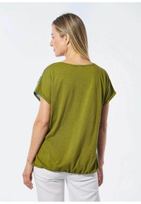 Grünes Kurzarmoberteil mit lockerem Schnitt, featuring einen gerafften Saum und gemusterten Ärmeln mit blau-weißen Streifen. Getragen mit weißen Hosen.