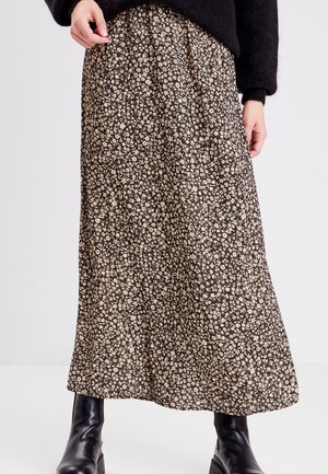 Vrouw draagt een zwarte trui, beige midi-rok met bloemenprint en zwarte leren enkellaarsjes, staand tegen een effen witte achtergrond.