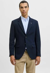 Blazer azul marino hecho de tejido texturizado, con dos bolsillos frontales, solapas con muescas y botones negros. Lleva puesto sobre una camisa de rayas azul claro.