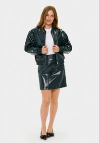 Giacca bomber in ecopelle lucida di un verde scuro, con polsini a costine, abbinata a una gonna mini coordinata. La modella tiene la giacca chiusa.