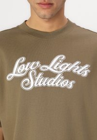 Oliven grøn t-shirt i bomuld med hvid broderet tekst, der står "Low Lights Studios." Klassisk crew neck design, blød tekstur.