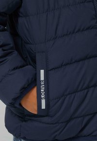 Marineblauwe pufferjack met een gequiltde textuur. Voorzien van een zijzak met een zilveren "Solver"-logo op het label. Het materiaal lijkt glanzend.