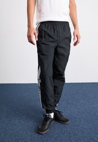 Černé lehké běžecké kalhoty s elastickým páskem, bílými postranními pruhy a logem. Nosené s černými teniskami a bílými ponožkami.