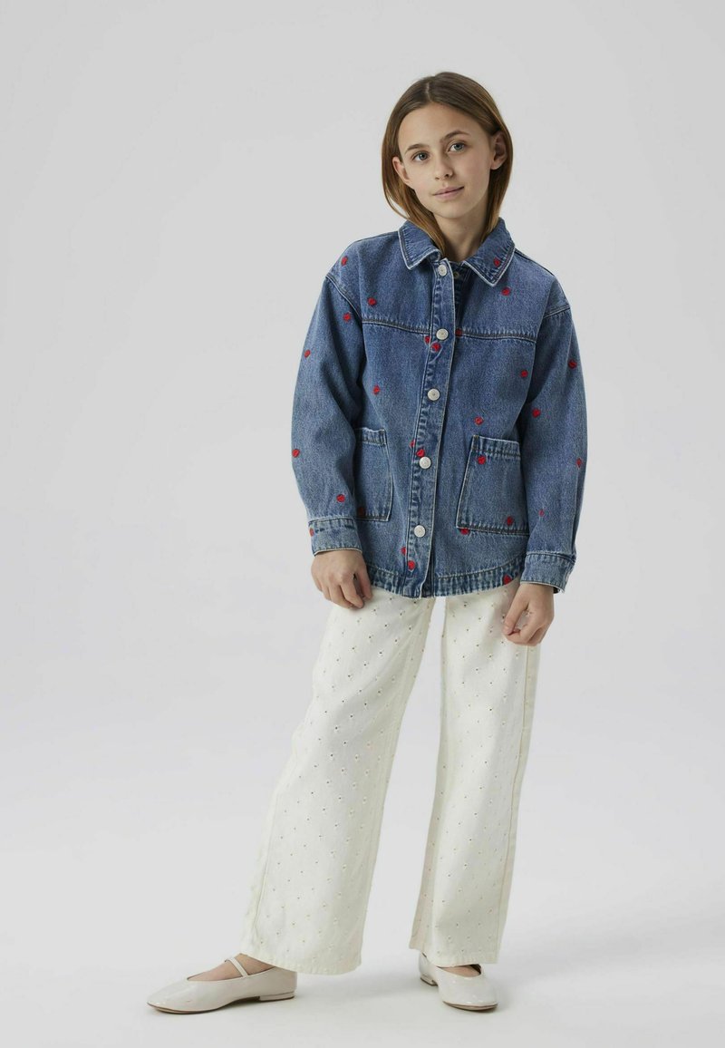 Veste en denim avec des cœurs rouges brodés, fermeture à boutons, deux poches avant, associée à un pantalon blanc à jambes larges et des chaussures plates.