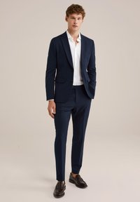 Costume bleu marine à coupe ajustée, blazer à bouton unique et pantalons sur mesure. Chemise blanche en dessous. Mocassins en cuir noir.