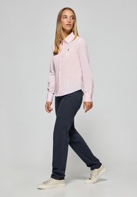  Lichtroze overhemd met lange mouwen, gecombineerd met donkerblauwe broek en beige sneakers. Het overhemd heeft een klein geborduurd logo.