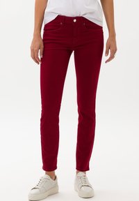 Jeans rojos ajustados con una textura suave, cinco bolsillos y cierre de botón, combinados con zapatillas blancas para un look casual.