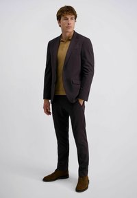 Mann steht mit einem dunklen Blazer, passenden Hosen, einem senfgelben Strick-Polo-Shirt und braunen Wildlederschuhen vor einem einfarbigen Hintergrund.