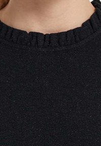 Nahaufnahme des Halses und oberen Brustbereichs einer Person, die einen schwarzen Pullover mit einem sanft gerafften, gerippten Kragen trägt.