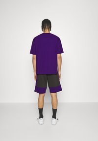 Fialové bavlněné tričko s krátkými rukávy, kombinované s černými a fialovými šortkami s kontrastním lemem. Bílými teniskami a černými ponožkami je vzhled dokončen.