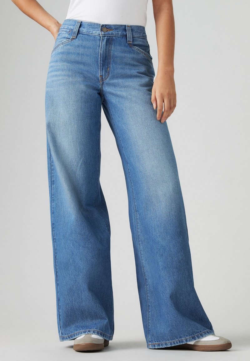 Personne portant un jean ample bleu clair et des baskets blanches, debout devant un fond gris clair uni.