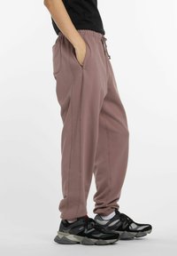 Persoon in mauve joggingbroek met zijzakken en zwart-grijze sneakers, staande tegen een witte achtergrond.