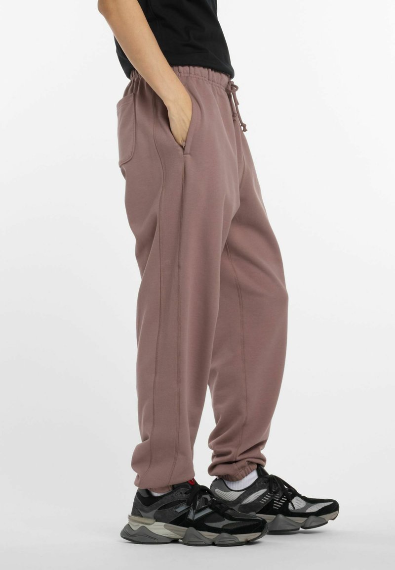 Persoon in mauve joggingbroek met zijzakken en zwart-grijze sneakers, staande tegen een witte achtergrond.