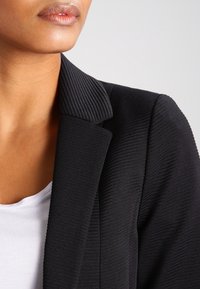 Nahaufnahme einer Person, die ein strukturiertes schwarzes Blazer über einem weißen Oberteil trägt. Der Fokus liegt auf dem Kragen und dem unteren Teil des Gesichts vor einem einfarbigen Hintergrund.