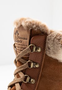 Botte en daim marron avec bordure en fausse fourrure, œillets en métal et lacets beiges. Présente un logo embossé et des détails cousus sur la pièce en cuir.