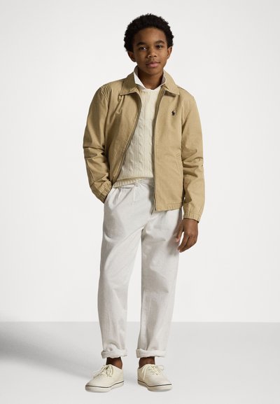 Khaki lettvektsjakke, kremfarget kabelstrikket genser, hvite bukser og lyse joggesko. Jakken har en liten logo. Teksturerte materialer.