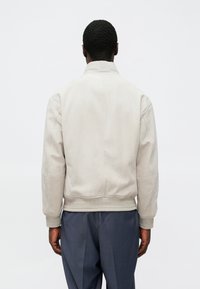 Veste bomber beige clair avec une texture lisse, col montant et poignets côtelés. Le dos présente un détail discret de couture. Portée avec un pantalon bleu foncé.