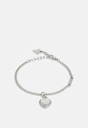 Silver chain bracelet with heart-shaped charm engraved "Guess" and a small triangular tag, featuring a lobster clasp closure.