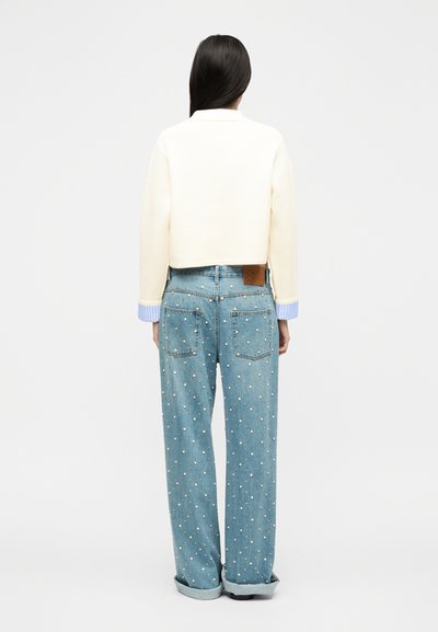 Femme debout, vue de dos, portant un pull court crème avec des poignets bleus et un jean large bleu clair orné de perles, retroussé à l'ourlet.