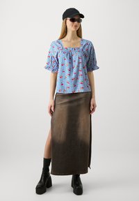 Blusa a quadri blu e bianca con stampe di ciliegie rosse, scollatura quadrata e maniche a sbuffo. Abbinata a una maxi gonna di denim marrone e stivali neri.
