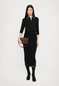 Schwarzer, figurbetonter Pullover mit blauem Kragen, kombiniert mit einem langen schwarzen Ripprock. Eine dunkelbraune Handtasche, schwarze Socken und Sandalen runden das Outfit ab.
