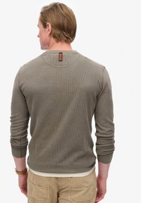 Man die een olijfgroen shirt met lange mouwen draagt met een klein label aan de bovenkant van de achterkant en beige broek, met de rug naar de camera gewend.