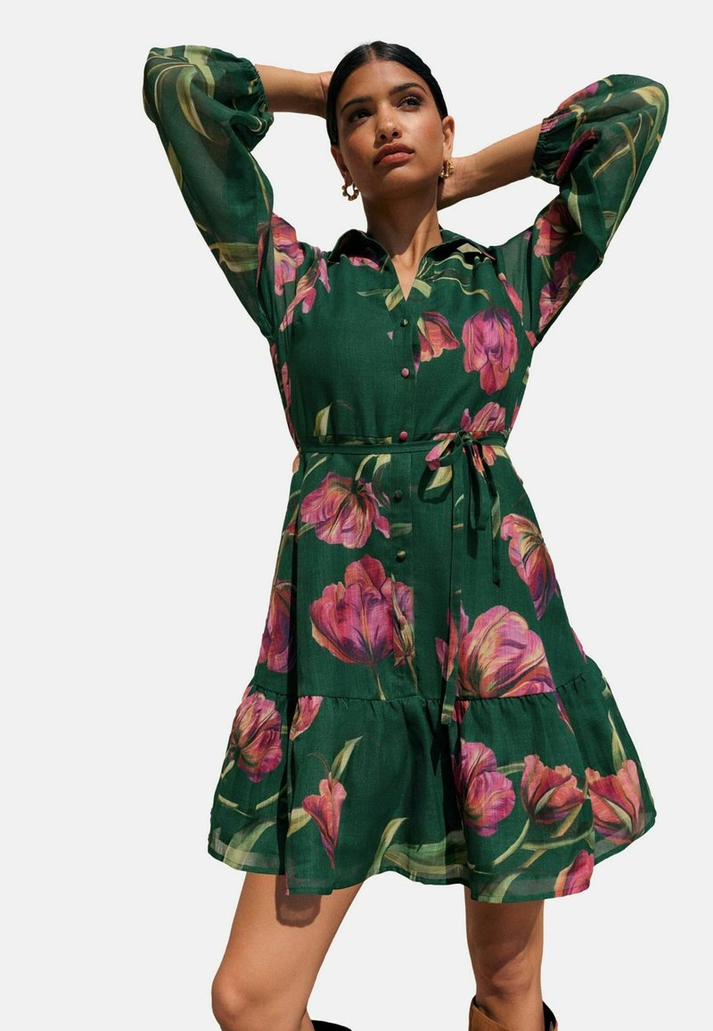 Žalia gėlių suknelė su rožinėmis tulpėmis, lengva medžiaga, sagų užsegimas priekyje, trumpi pūsti rankovės, juosmens virvė ir sluoksniuota apačia.