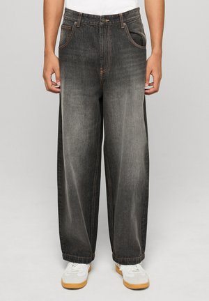 Jeans denim grigi scuro, taglio largo, con trama sbiadita, cuciture arancioni, cinque tasche e chiusura con bottone, abbinati a sneakers bianche.