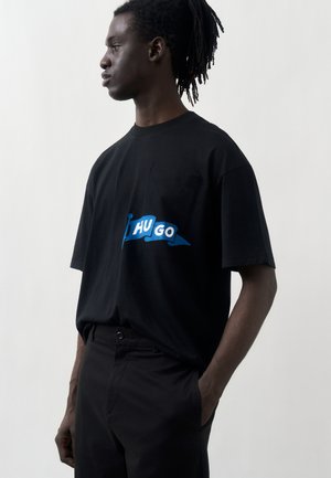 Sötét bőrű, raszta hajú férfi fekete nadrágot és fekete pólót visel, amelyen kék „HUGO” zászlólogó található, oldalról látható, egyszínű háttér előtt áll.