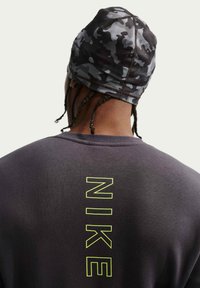 Άτομο που φοράει μαύρο κάλυμμα κεφαλής με καμουφλάζ και σκούρο πουκάμισο με κάθετο κείμενο "NIKE" με κίτρινο περίγραμμα στην πλάτη.