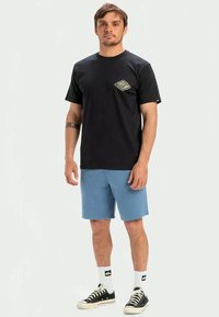 Camiseta de algodón negra con un pequeño gráfico colorido en el pecho, combinada con pantalones cortos azul claro, zapatillas negras y calcetines blancos.