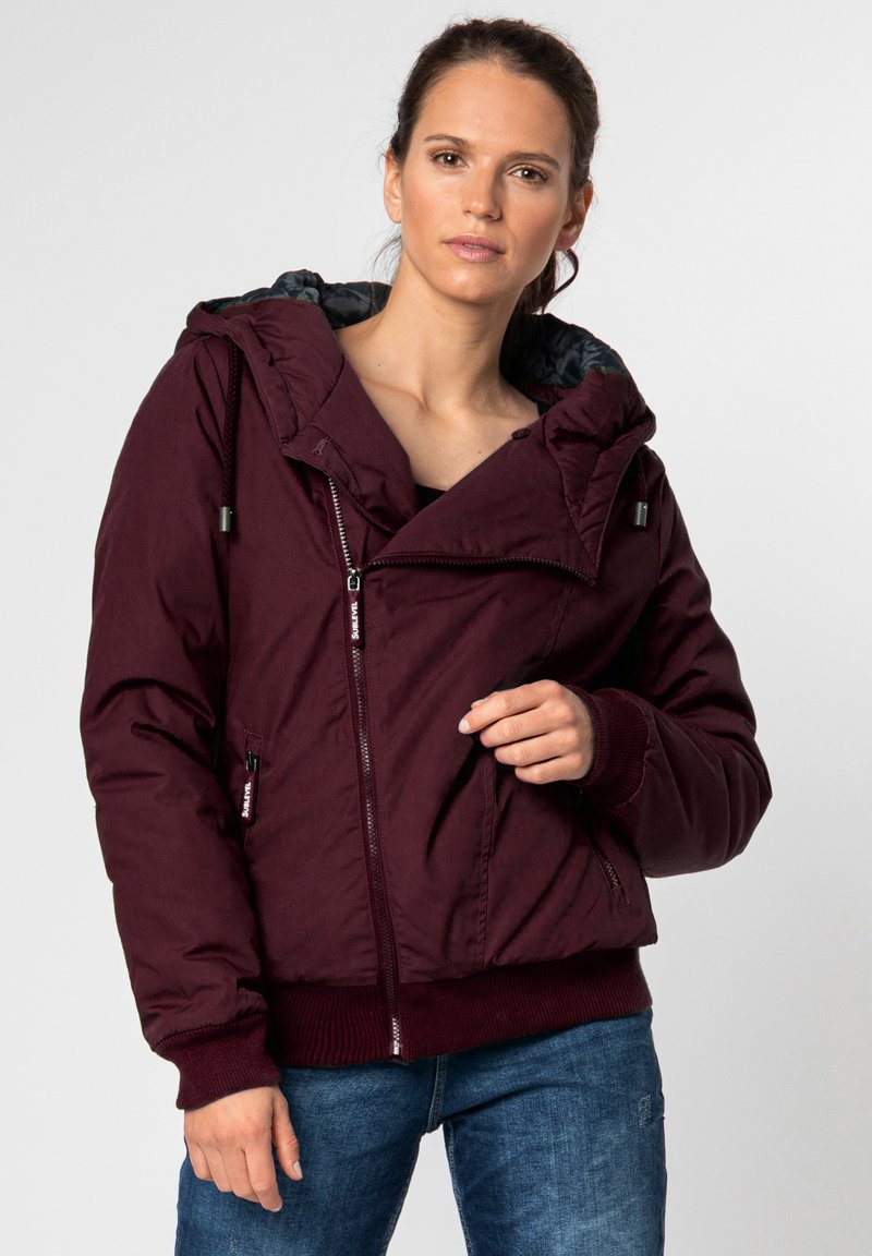 Burgundy puffer jacket with large hood, zippered front, and ribbed cuffs. Features side pockets and a smooth, textured exterior.