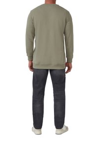 Uomo che indossa un felpa a maniche lunghe verde oliva, jeans grigio scuro e scarpe da ginnastica bianche, in piedi di spalle contro uno sfondo bianco.