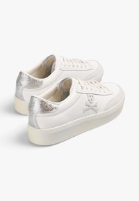 Zapatillas bajas blancas con tabiques de talón plateados y un diseño bordado de calavera y huesos cruzados en el costado, con cordones y suelas gruesas.