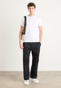 Witte t-shirt met korte mouwen, zwarte wijde broek, witte sneakers. Model houdt een beige tas met een zwarte strap vast, minimalistisch ontwerp, effen oppervlakken.