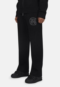 Czarne dresowe spodnie z dzianiny z prążkowanymi mankietami, z szarym wzorem logo na lewej udzie. Dobrane z czarnymi trampkami, tworząc spójną całość.