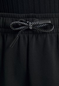 Cintura de tecido preto com material elástico, apresentando uma faixa elástica plana e um cordão atado com costura em zig-zag e extremidades de plástico.