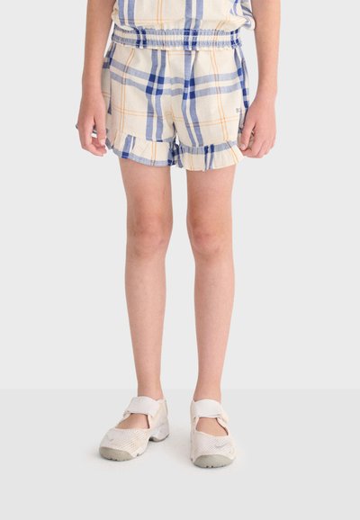 Enfant debout portant un short crème à carreaux bleus et orange, avec des bords froncés, et des chaussures blanches en mesh avec des brides.