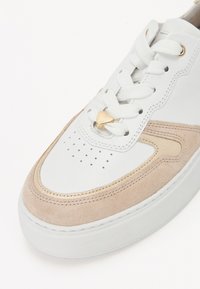 Baskets blanches et beiges avec un bout perforé, lacets blancs passés dans des œillets dorés et un petit pendentif en forme de cœur doré sur les lacets.
