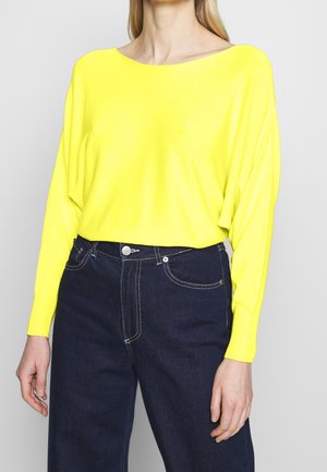Ljusgul stickad tröja med bred båtutskärning och långa ärmar, kombinerad med mörkblå högmidjade jeans med kontrasterande sömmar.